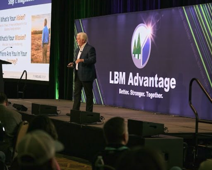 A speaker addressing an audience from an LBM Advantage stage, with the Better. Stronger. Together. backdrop behind him and a presentation slide to one side